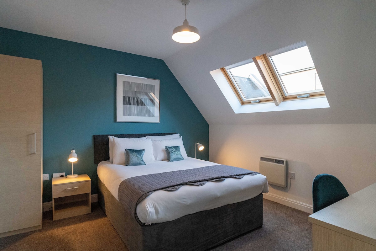 A bedroom with a bed, bedside tables, a desk, a chair, a window, and a ceiling light.