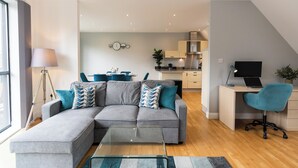 Un apartament modern amb un sofà seccional gris, una taula de centre de vidre, un escriptori amb un ordinador portàtil i una cadira de despatx blava.