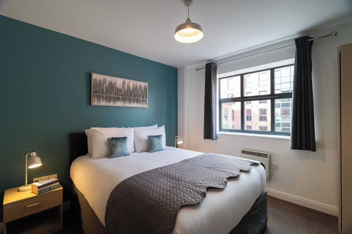 A neatly arranged hotel room with a large bed, a bedside table with a lamp, a window with curtains, and a wall-mounted artwork.