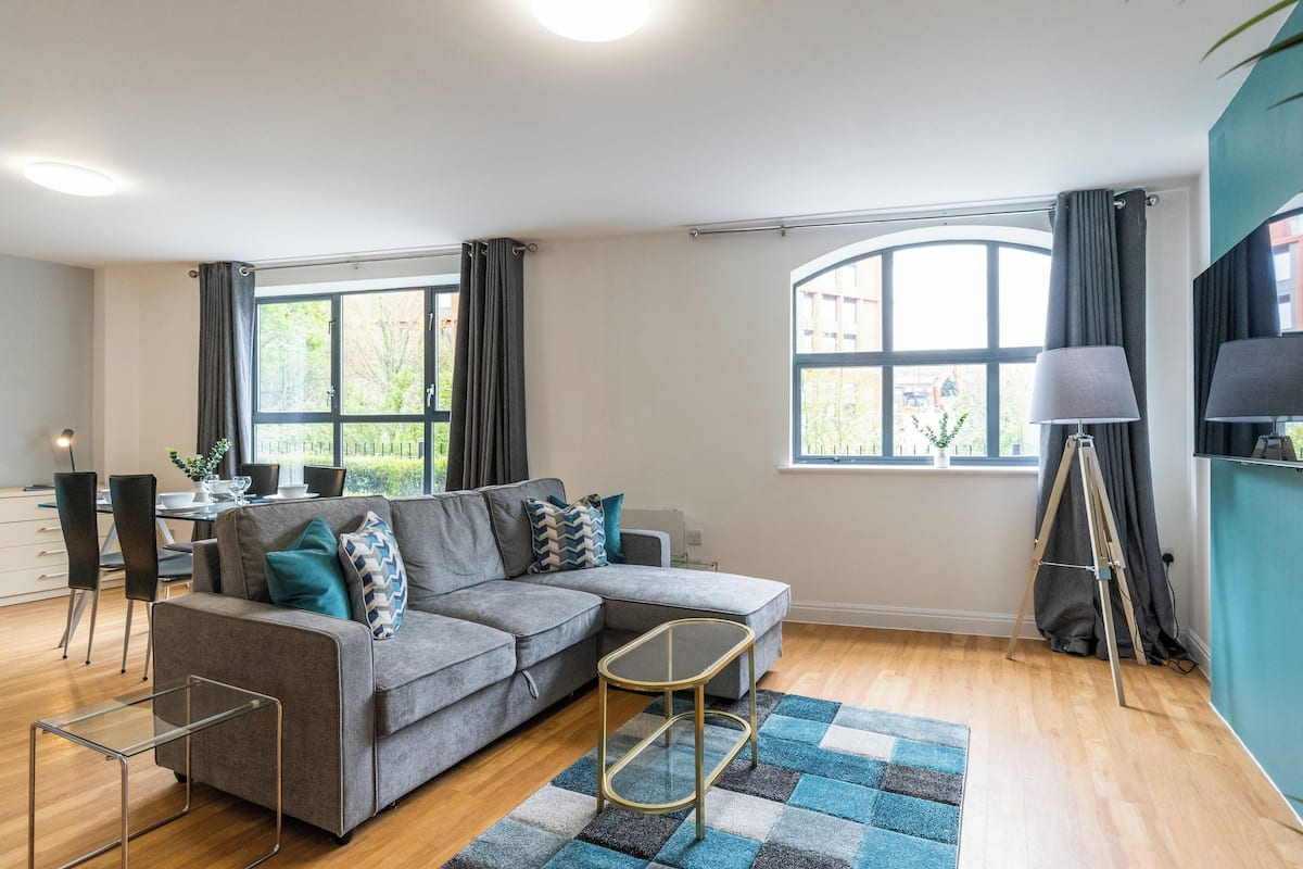 A modern living room with a grey sectional sofa, a small glass coffee table, a tripod floor lamp, and a large window with curtains.