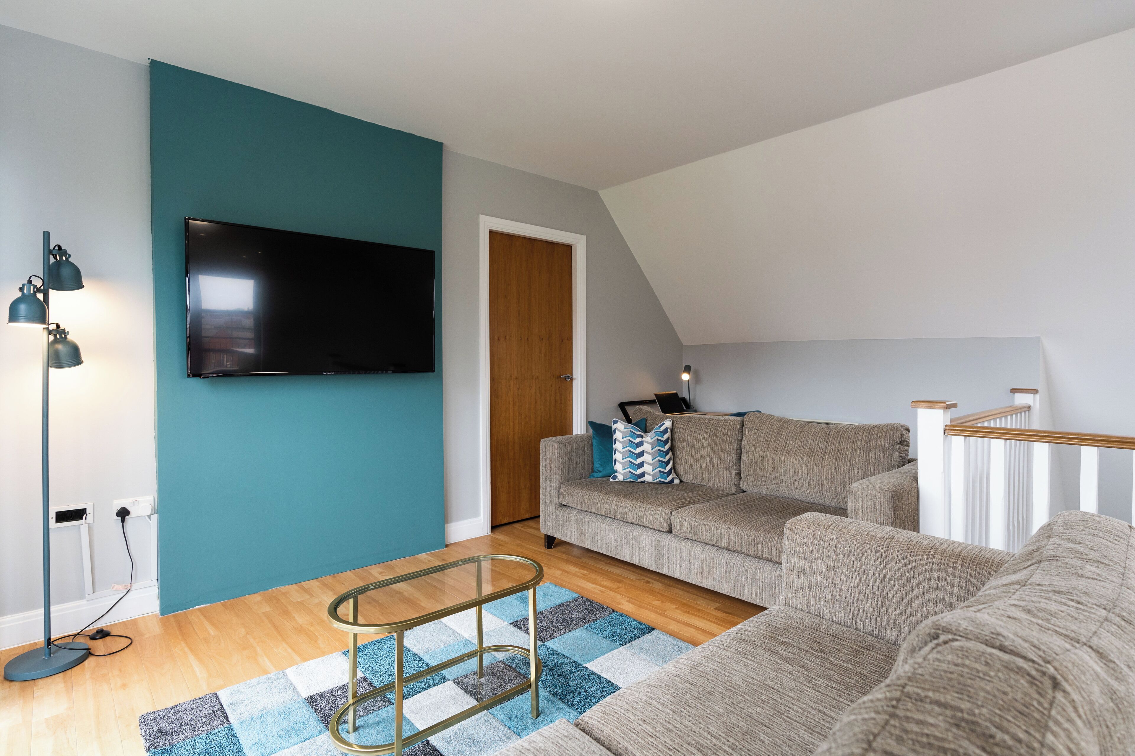 A modern living room with a teal accent wall, a flat-screen TV, a sofa, a coffee table, and a staircase.