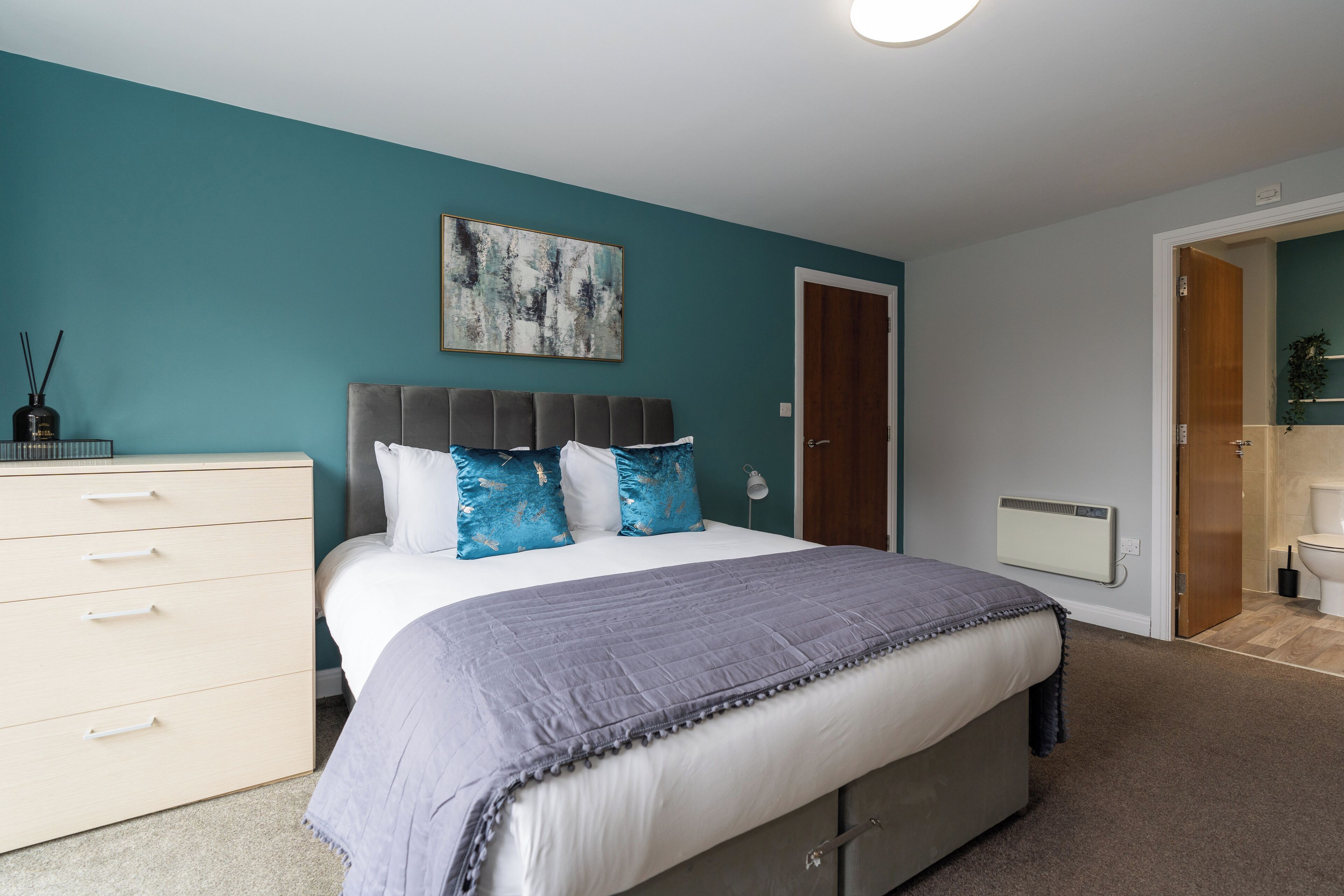 A bedroom with a bed, a dresser, a painting, and a bathroom door.