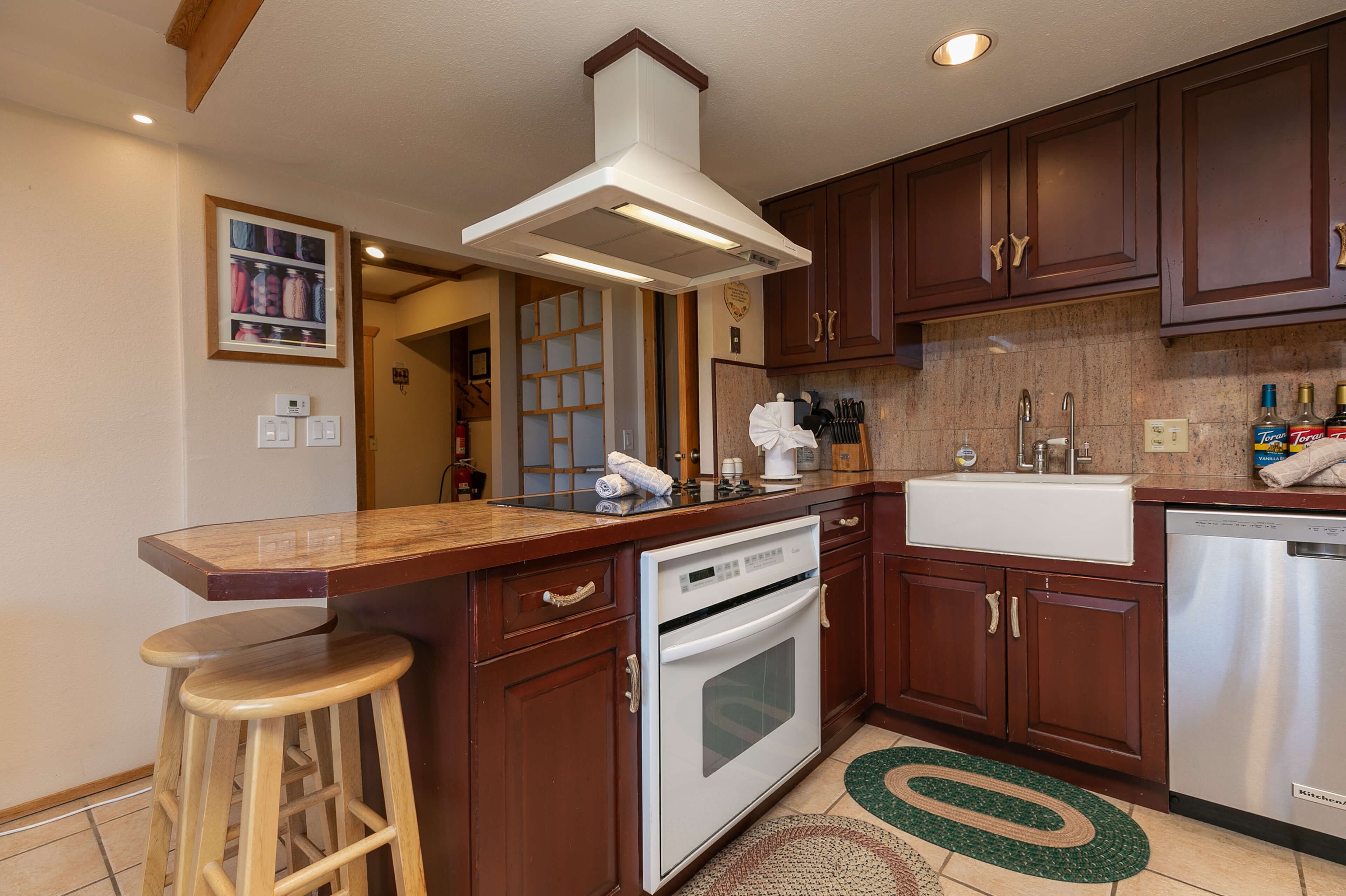 A kitchen with wooden cabinets, a stainless steel dishwasher, a built-in oven, a countertop with a sink, and a bar stool.