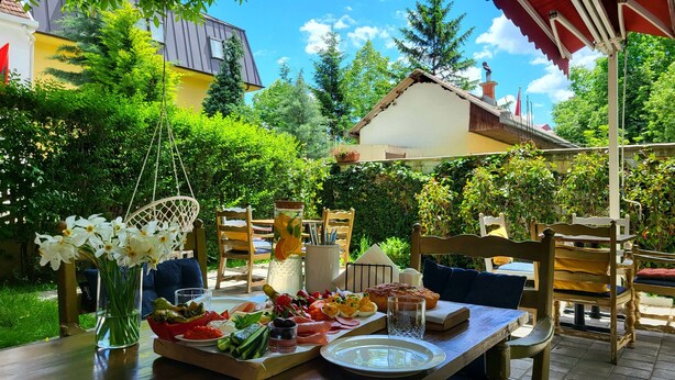 Un área de comedor al aire libre con una mesa preparada para una comida, rodeada de exuberante vegetación y un edificio al fondo.