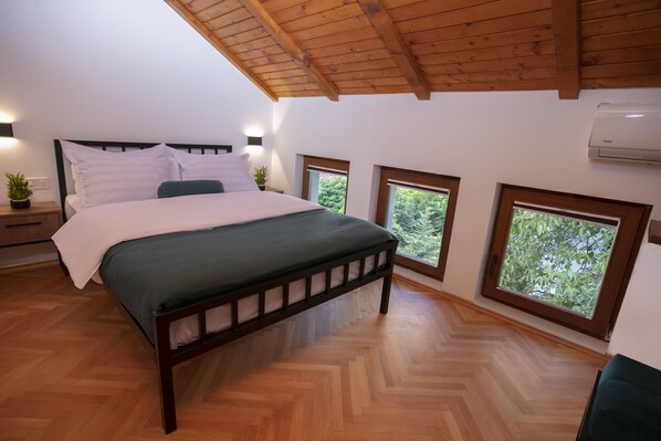 A bedroom with a wooden bed, a green mattress, a wooden headboard, a nightstand with a plant, and two windows with a view of trees. - Grand Boutique Hotel (Pristina)