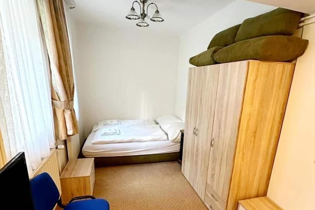 A small, compact room with a single bed, a wardrobe, a desk with a chair, and a ceiling light fixture.