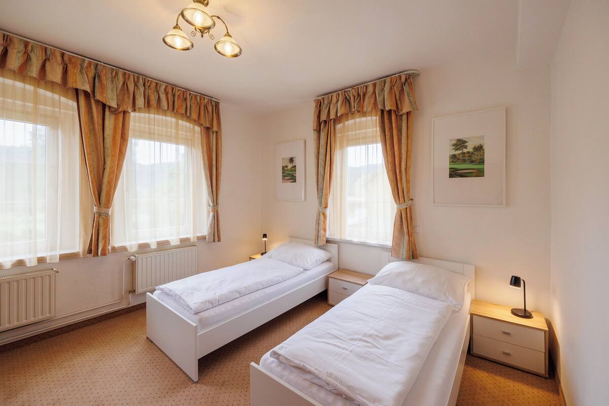 A twin room with two single beds, a bedside table with a lamp, a radiator, and a window with curtains.