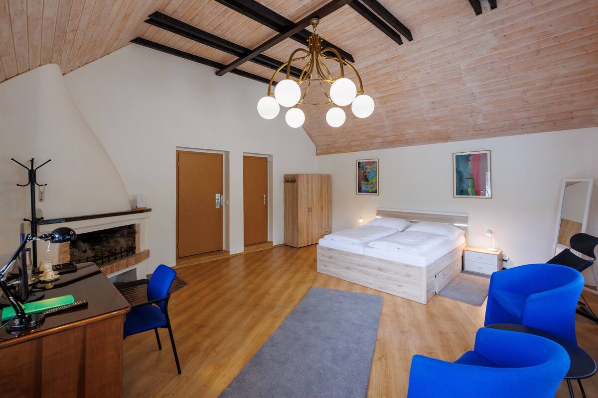 A modern bedroom with a loft-style ceiling, a wooden desk with a lamp, a bed with a white mattress, and two blue chairs.