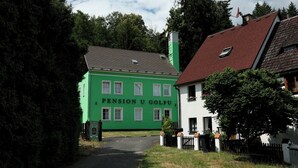 Un edificio verde con techo rojo, identificado como "Pension U Golf", rodeado de árboles y un edificio blanco con una valla negra.