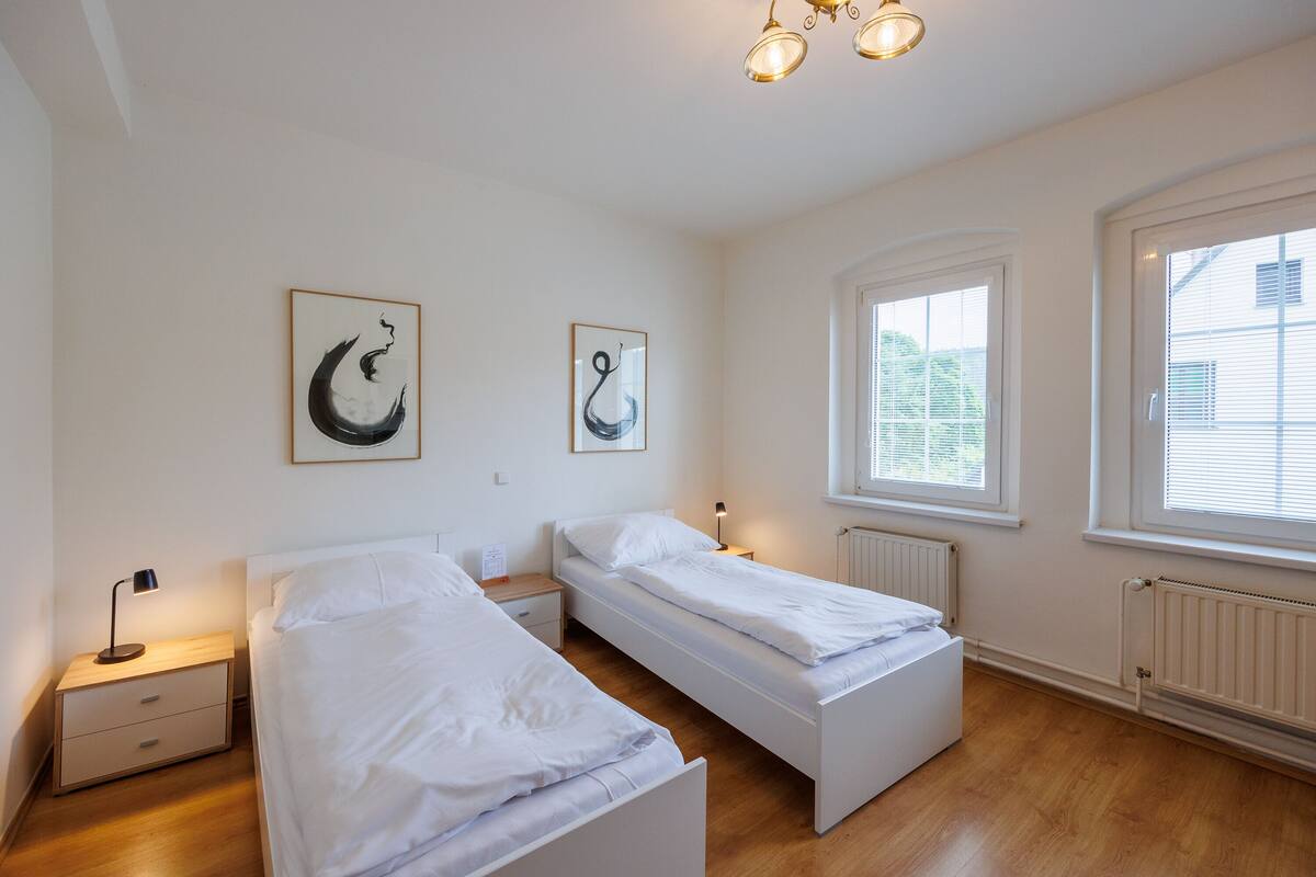 A bedroom with two single beds, a nightstand with a lamp, a window with blinds, and artwork on the wall.