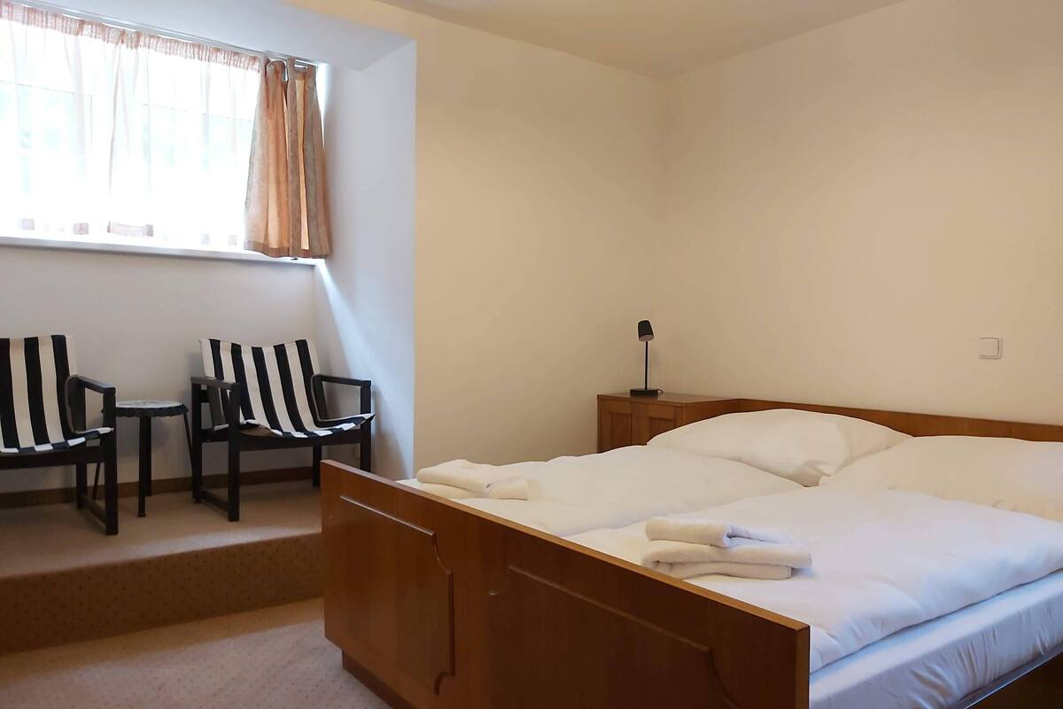 A bedroom with a wooden bed, a small bedside table with a lamp, two striped chairs, and a window with curtains.