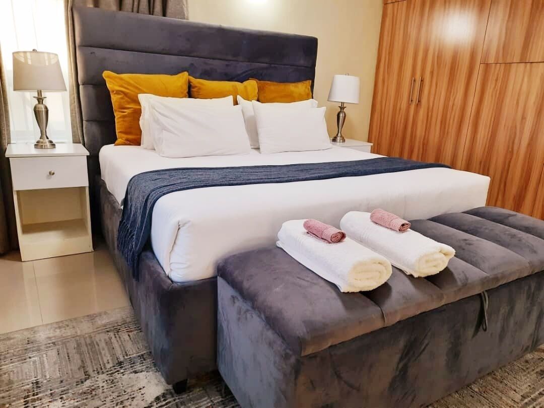 A neatly made bed with a dark headboard, white and yellow pillows, and a grey ottoman with towels.