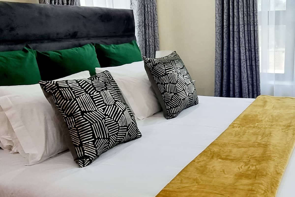 Uma cama bem arrumada com almofadas brancas e estampadas, um cobertor dourado e um conjunto de toalhas sobre um sofá de cor escura.