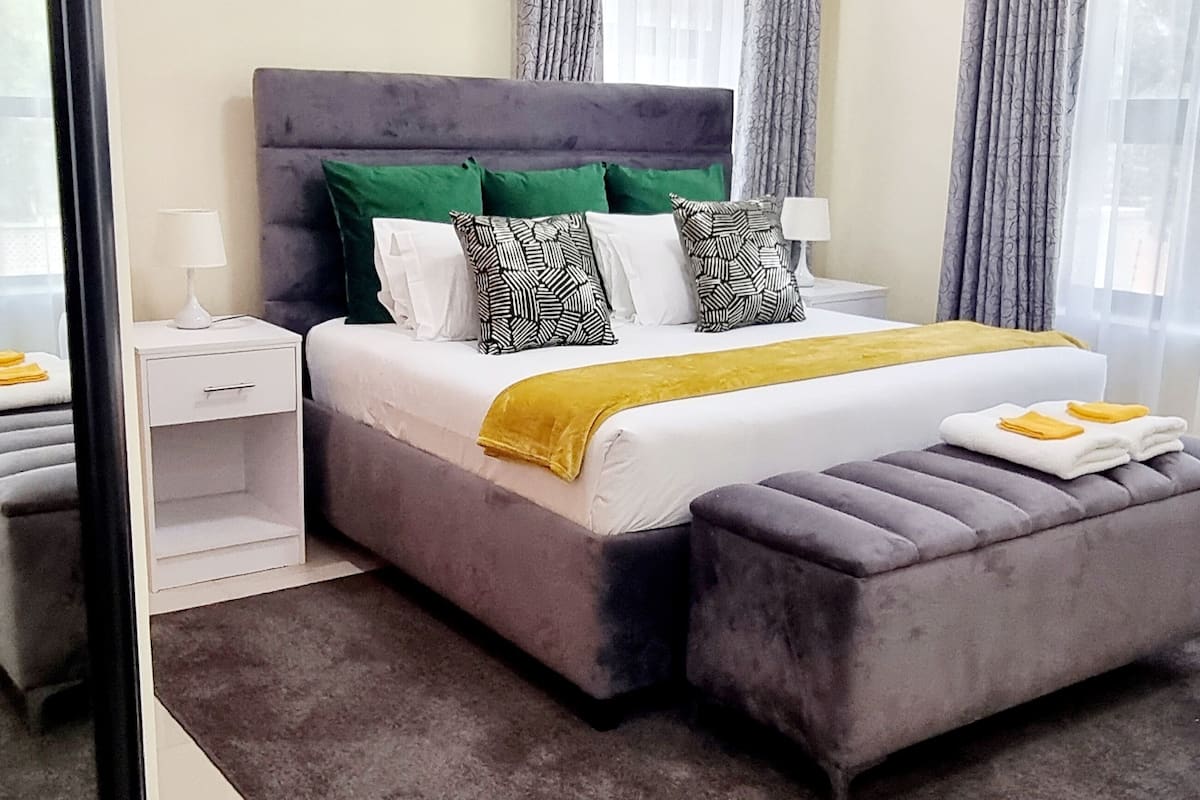 Quarto de hotel moderno com uma cama grande, mesa de cabeceira, um banquinho cinza e uma janela com cortinas finas.