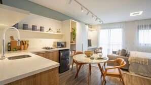 A modern kitchen with a dining area, featuring a built-in oven, microwave, and sink. There is a small refrigerator, wooden chairs, and a round dining table.