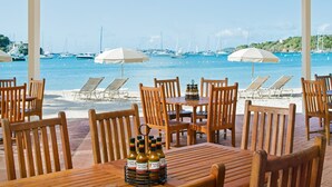 Un restaurant situé en bord de mer, avec des tables et des chaises en bois, une table où sont posées des bouteilles de bière, et une vue sur les bateaux et la mer.