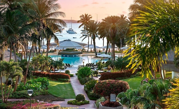 A resort with a central pool, surrounded by palm trees and a view of boats on the water.