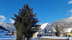 Un paisaje nevado con un gran pino en primer plano, un edificio blanco de techo inclinado y una montaña al fondo.