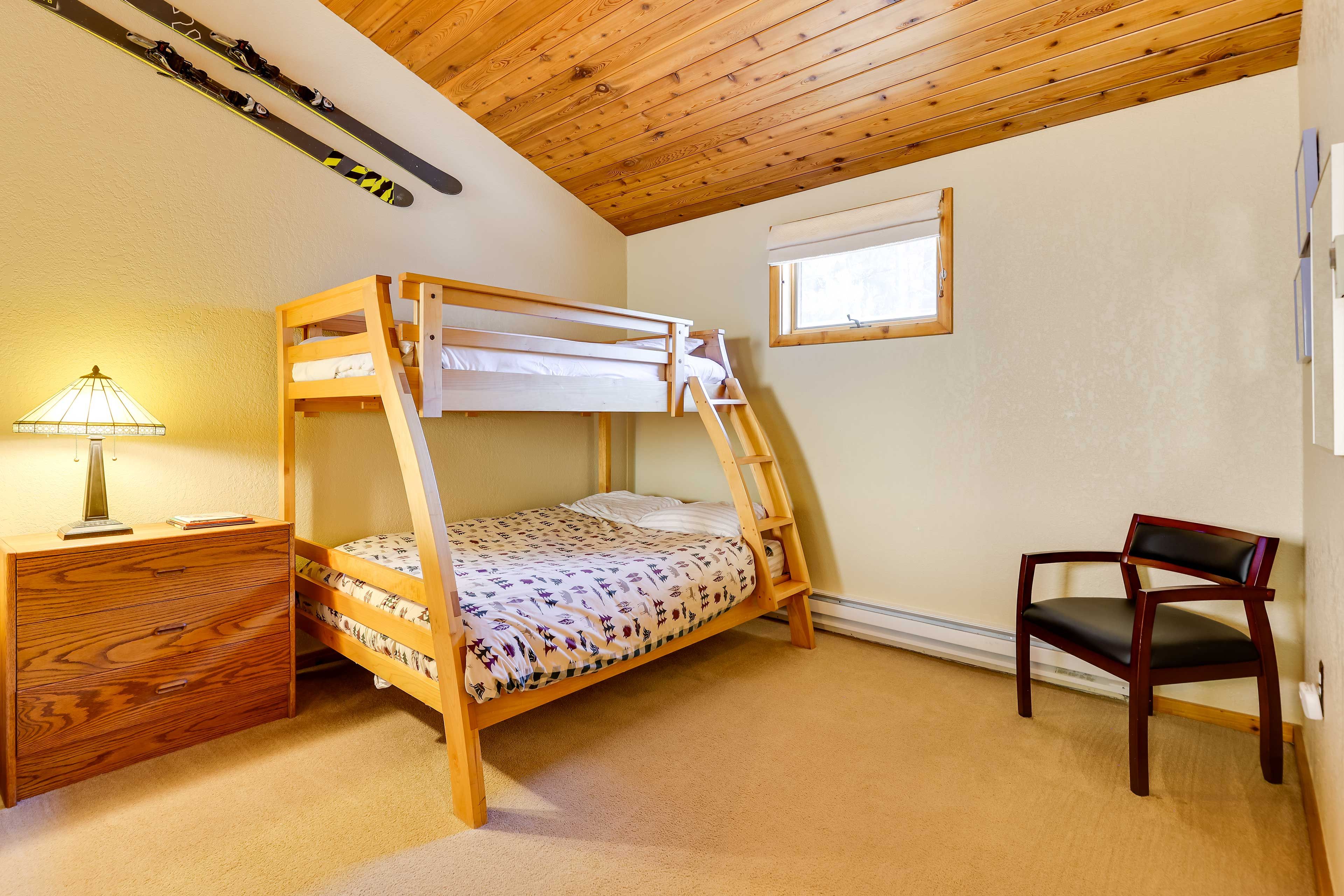 A bunk bed with a wooden ladder, a desk with a lamp, a chair, and skis hanging on the wall.