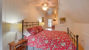 Une chambre avec un plafond en bois, un lit recouvert d’une courtepointe à motifs floraux rouges, une table de chevet avec une lampe et une fenêtre donnant sur l’extérieur.