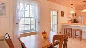 Une table à manger en bois, sur laquelle se trouve un saladier rempli de fruits, des chaises en bois, une porte blanche et un coin cuisine avec des tabourets et des appareils électroménagers.