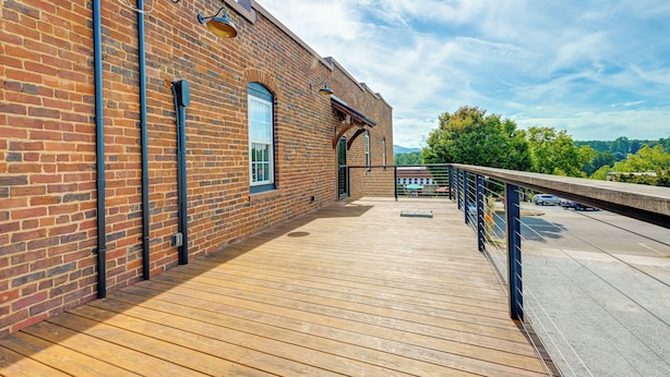 Une terrasse en bois, un bâtiment en briques, une rambarde en métal et un ciel dégagé.