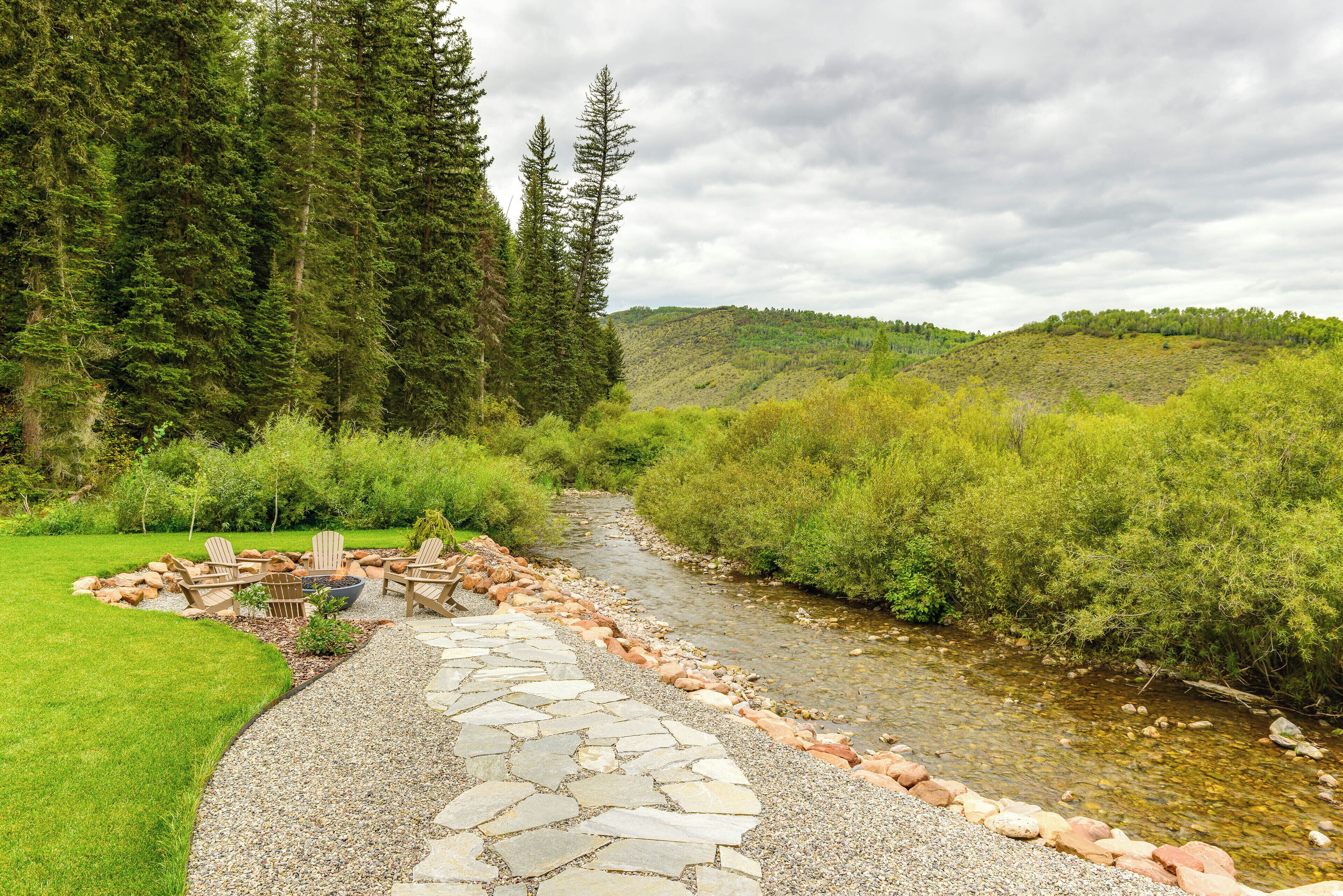 A stone pathway leading to a seating area with chairs and a fire pit, surrounded by lush greenery and a stream.