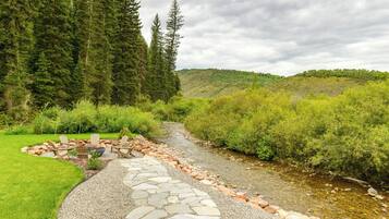 A stone pathway leading to a seating area with chairs and a fire pit, surrounded by lush greenery and a stream.