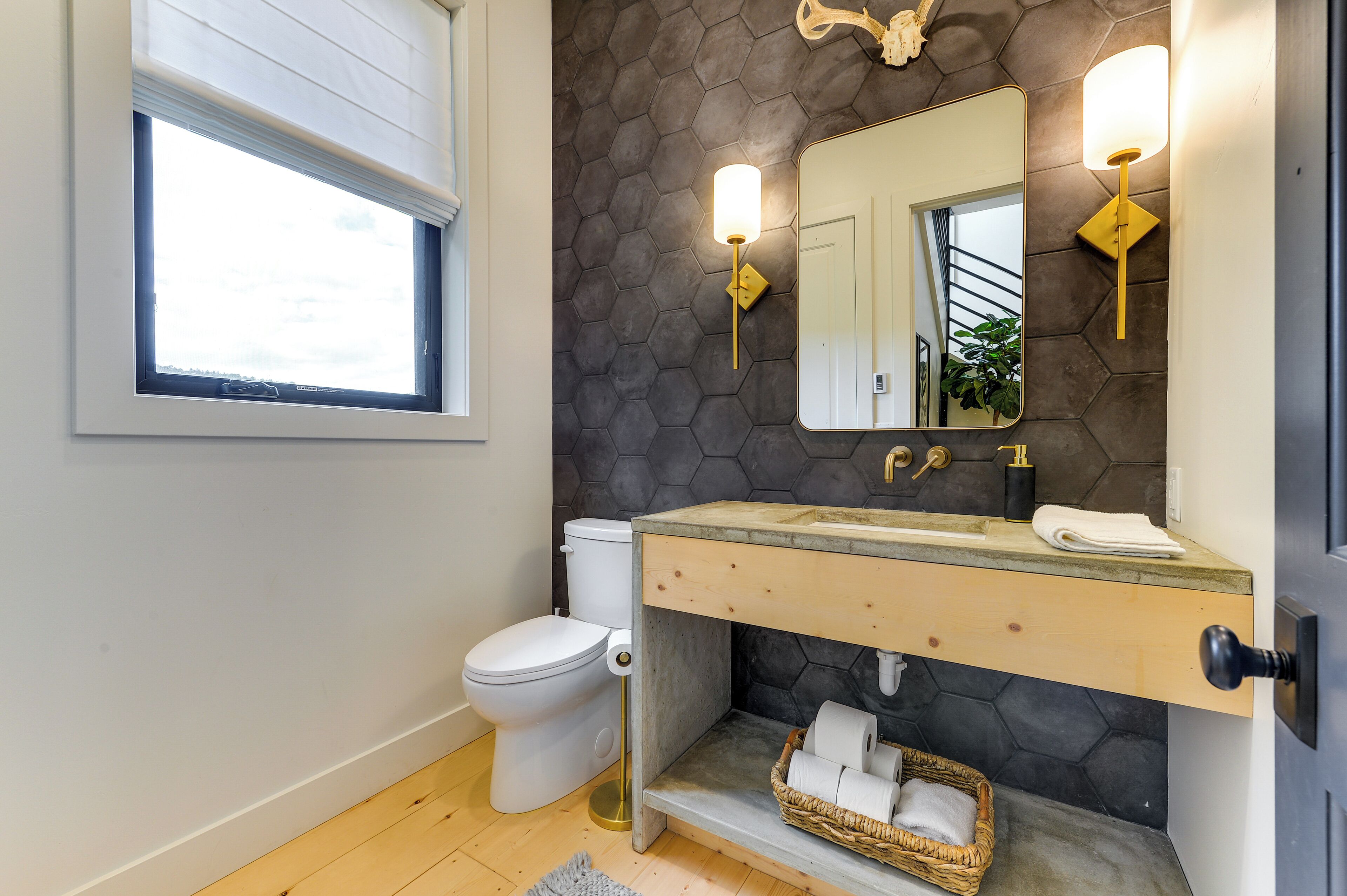 A modern bathroom with a hexagonal tile wall, a white toilet, a wooden vanity, a large mirror, and a window with blinds.