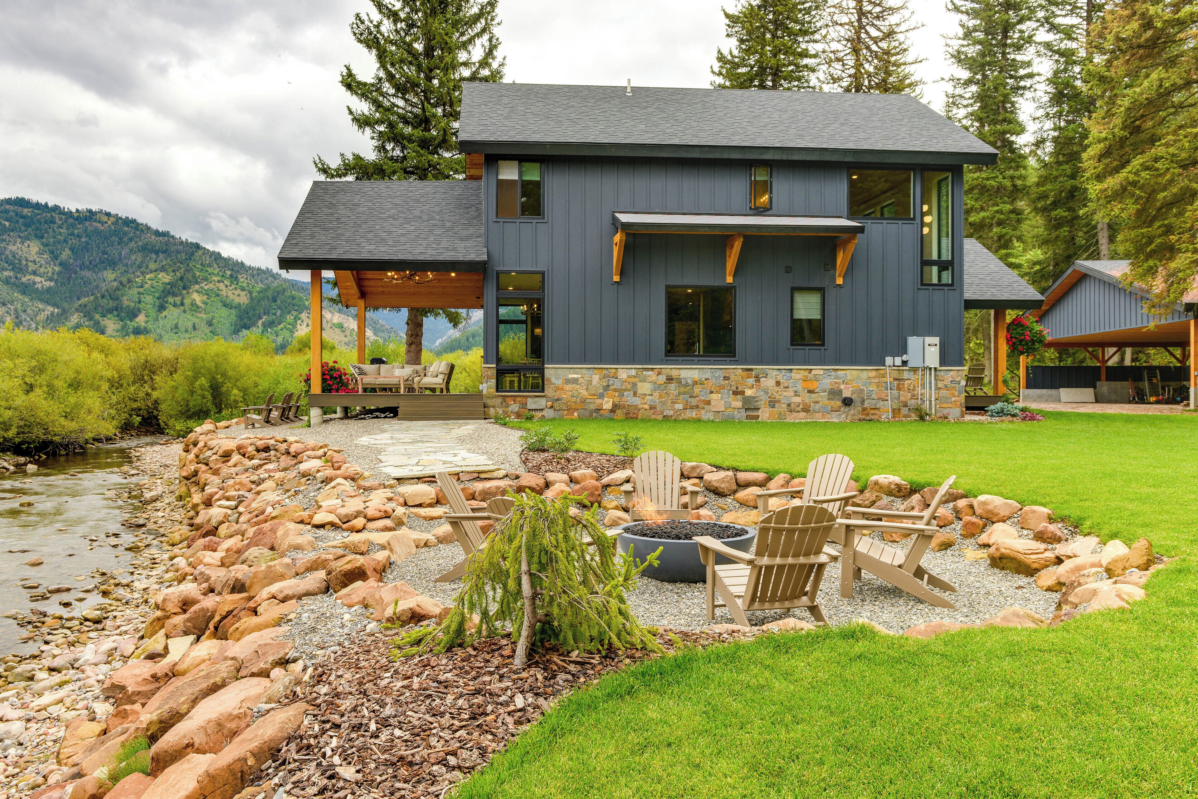 Une maison moderne avec des fondations en pierre, un patio doté de fauteuils Adirondack et une vue sur une rivière et des montagnes.