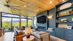 A modern living room with a fireplace, large windows, and a ceiling fan. - Modern Turnerville Cabin w/ Hot Tub & Scenic Views (Bedford)