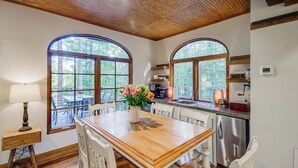A dining area with a wooden table, white chairs, and a vase of flowers. There are large arched windows with a view of trees. - Mississippi Retreat w/ Hot Tub, Deck & Lake Views! (Terry)