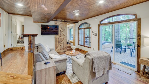 A spacious living room with a wooden ceiling, a large window, and a sliding glass door leading to an outdoor deck. - Mississippi Retreat w/ Hot Tub, Deck & Lake Views! (Terry)