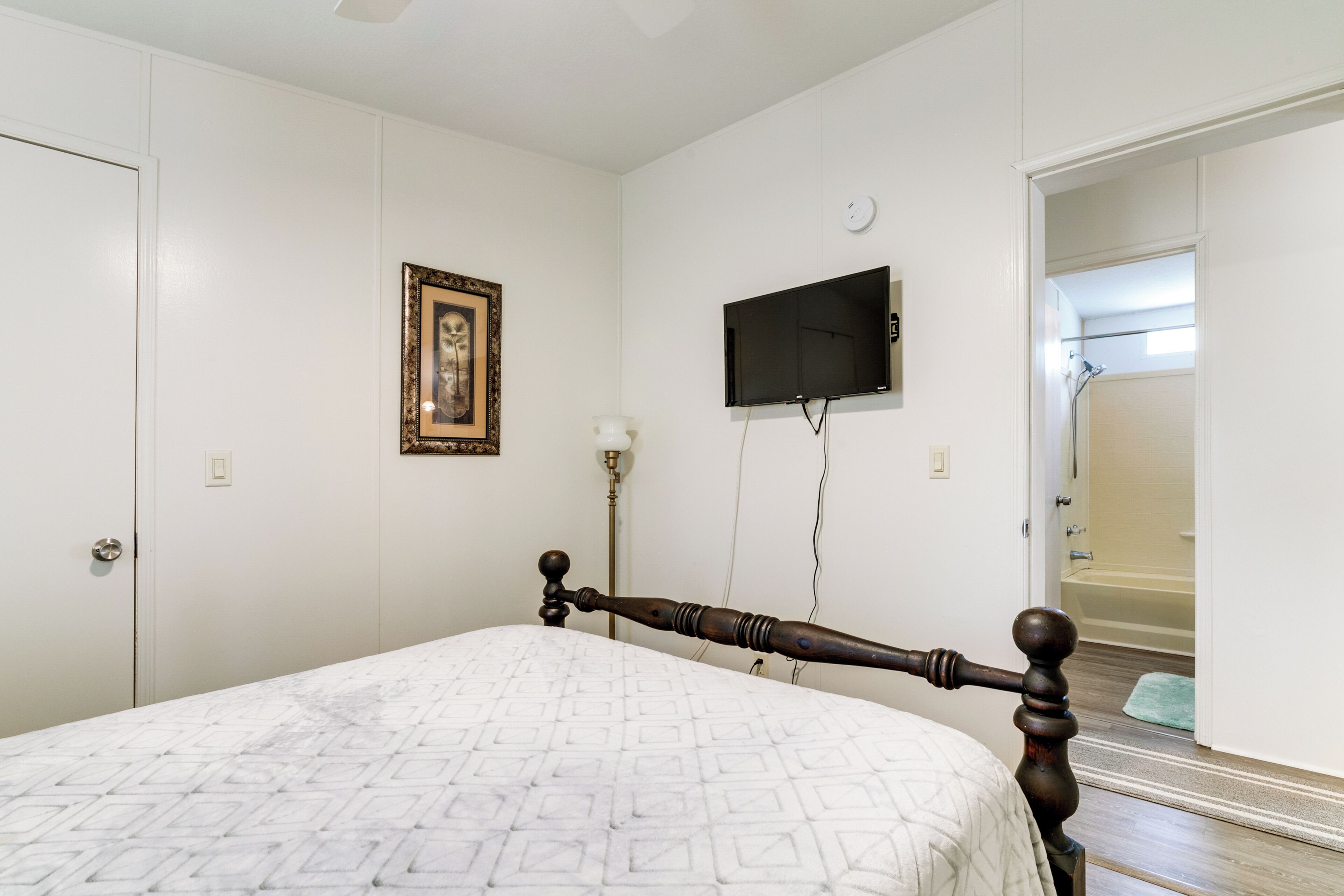 A bedroom with a bed, a television mounted on the wall, a framed picture, and a door leading to a bathroom.
