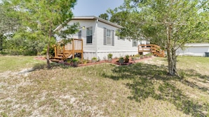 A single-story house with a wooden deck, surrounded by a landscaped garden with various plants and a small tree. - Weeki Wachee Home w/ Fire Pit: 13 Mi to Ocean! (Brooksville)