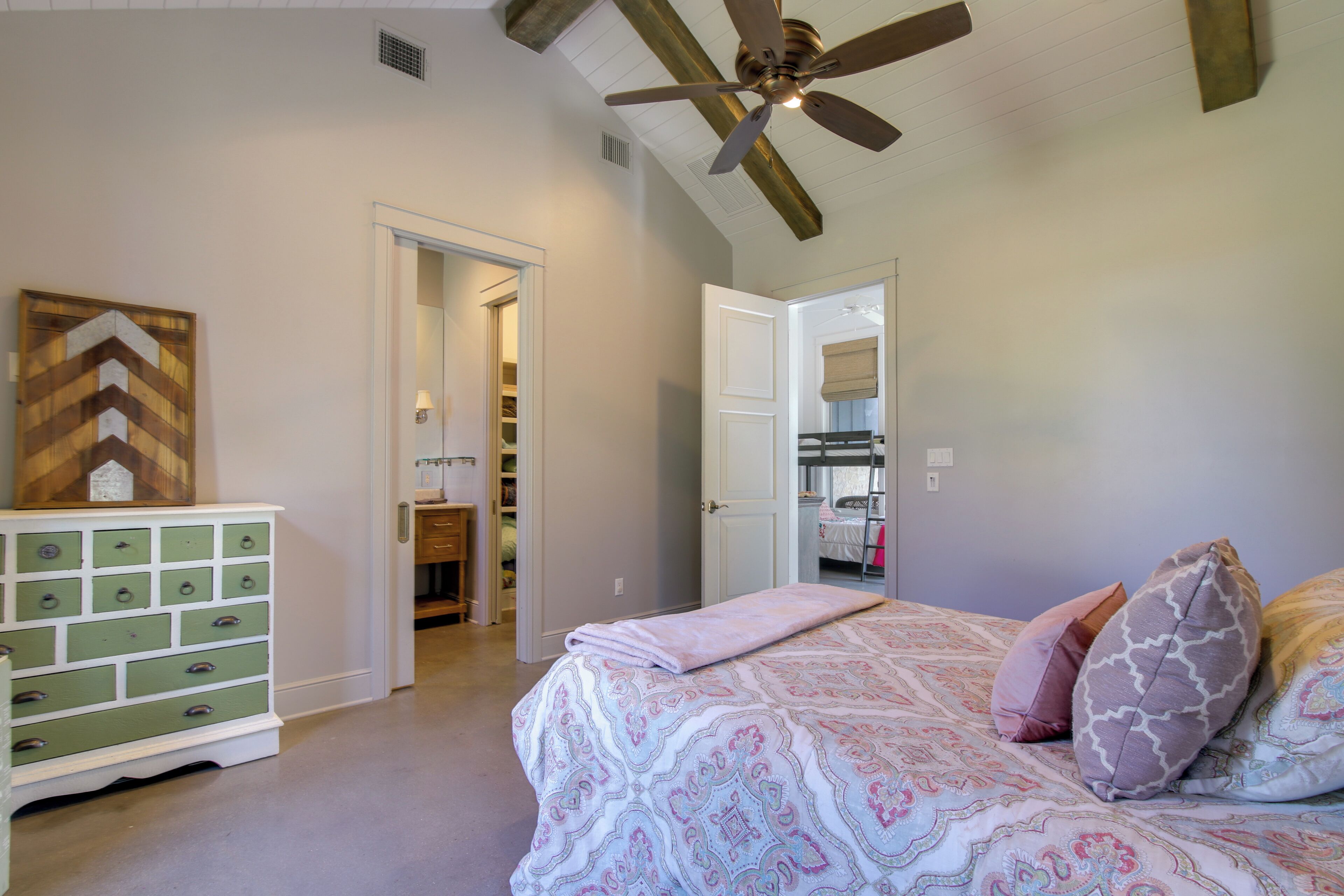 Un dormitorio con cama, ventilador de techo, cómoda y una puerta que da a un baño.