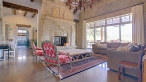 A spacious living room with a stone fireplace, a dining area with red upholstered chairs, and a large window providing natural light. - Pool & Covered Patios: Sprawling Austin Estate! (Austin)