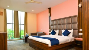 A hotel room with a large bed, a desk, a chair, and a view of greenery through the window. - HITE Resort Greater Noida (Greater Noida)
