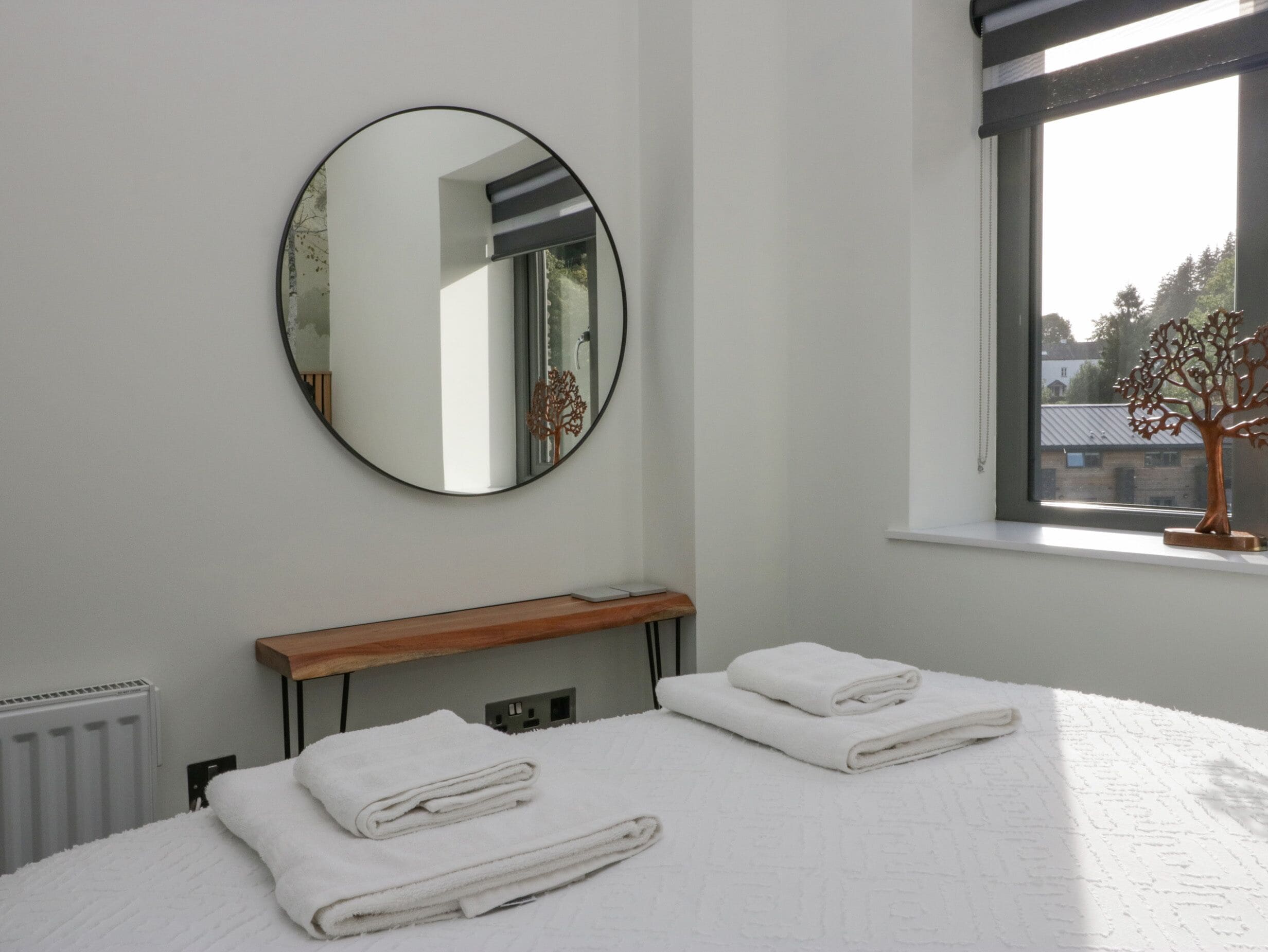 Camera d'albergo con un letto, uno specchio rotondo, una mensola in legno e una finestra vista edifici e alberi.