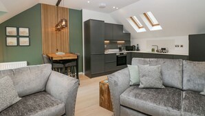 A modern living room with grey sofas, a wooden dining table, and a kitchen with dark cabinets and a skylight. - Watch Tree Apartment (Ulverston)