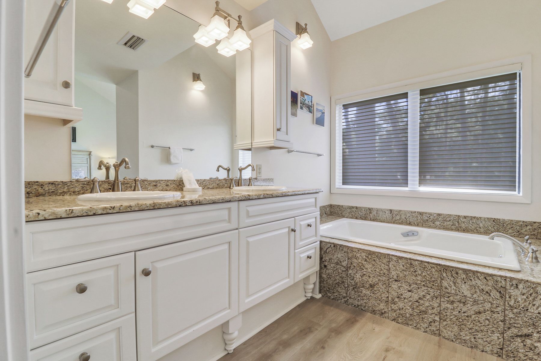 A bathroom with two sinks, a large mirror, a vanity with drawers, a bathtub, and a window with blinds.