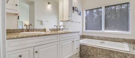 A bathroom with two sinks, a large mirror, a vanity with drawers, a bathtub, and a window with blinds.