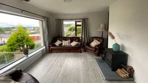 Uma sala de estar ampla com uma grande janela, um sofá de couro marrom, piso de madeira e um rack preto para TV.