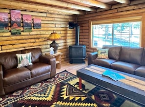 L’intérieur d’une cabane en rondins, avec deux canapés en cuir, une table centrale, une cheminée et une fenêtre donnant sur la forêt.