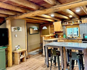 L’intérieur d’un chalet rustique présente des poutres apparentes, un coin cuisine avec des meubles en bois, un bar avec des tabourets et un téléviseur.