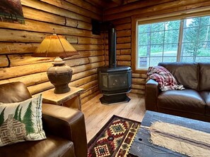 L’intérieur d’une cabane en rondins, avec un poêle à bois, des meubles en cuir et une fenêtre donnant sur la forêt.