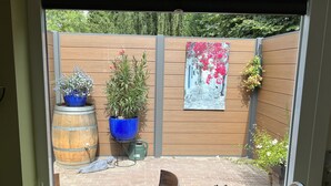 A window view of a backyard with a wooden fence, potted plants, a painting, and a brick patio.
