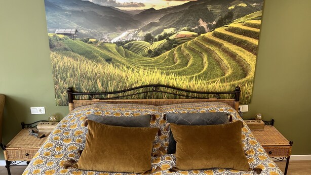 A bedroom with a large landscape painting of terraced fields and mountains, a bed with patterned bedding, and two pillows.