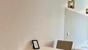 Una habitación minimalista con una mesa blanca y dos sillas, una cama con ropa blanca y dos apliques de pared.