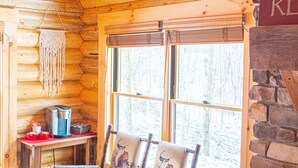 L’intérieur d’un chalet confortable, avec des murs en rondins, un plafond en bois, une cheminée en pierre, une fenêtre donnant sur la forêt et une table sur laquelle se trouvent une machine à café et une chaise ornée de motifs d’élans.